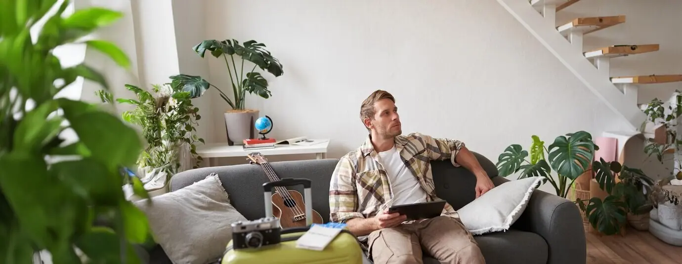 Porträt eines gutaussehenden jungen männlichen Touristen, der mit einem digitalen Tablet neben einem Koffer und zwei Tickets sitzt, online etwas für seinen Urlaub bucht und sich auf die Ferien vorbereitet.